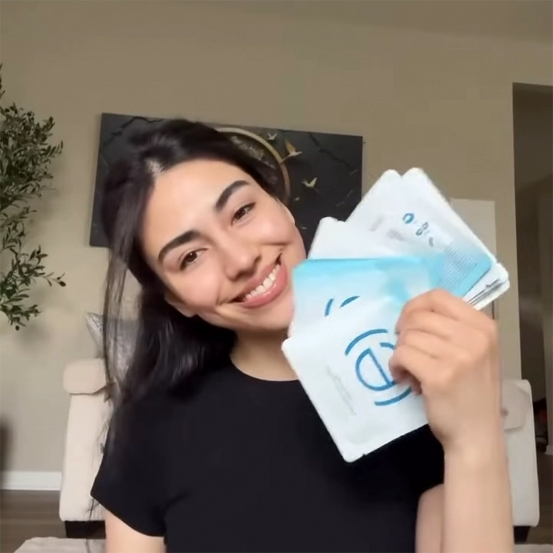 Woman holding a stack of white packets of In My Eyes Eye Patches / Eye Mask with blue designs in a home setting