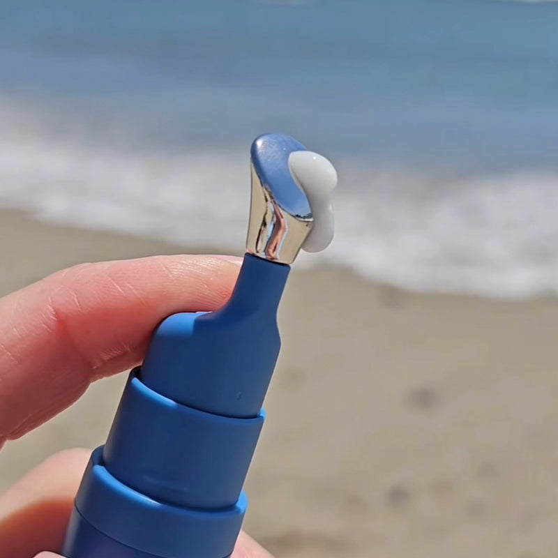 Blue lip balm with a silver cap held by a hand on a beach. In My Eyes Eye Serum, Zamak Pump