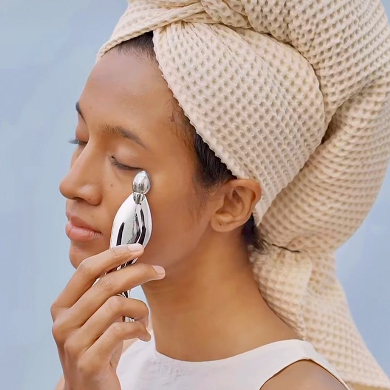 Woman applying skincare product with In My Eyes Mermaid Tail Gua Sha Roller against a light blue background